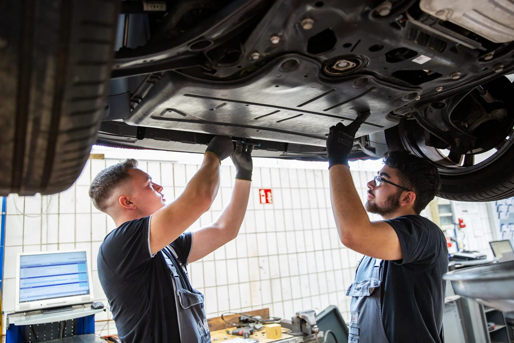Zwei Kfz-Mechatroniker arbeiten unter einem Auto in einer Werkstatt.